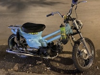 A vintage motorbike with light blue body paint and a single seat. It features a rugged off-road tire at the front and a metal luggage rack at the back. A plastic water bottle is attached to the frame near the handlebars. The surroundings include a road surface with scattered leaves.