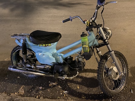 A vintage motorbike with light blue body paint and a single seat. It features a rugged off-road tire at the front and a metal luggage rack at the back. A plastic water bottle is attached to the frame near the handlebars. The surroundings include a road surface with scattered leaves.