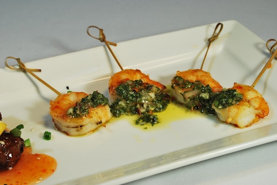 Grilled shrimp skewers garnished with a green herb sauce are arranged neatly on a white rectangular plate. The presentation is elegant, with drizzled sauce adding a touch of color and flavor. There is also a small portion of another dish in the corner with diced vegetables and sauce.