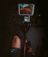A person is riding a stationary bike indoors, wearing cycling shorts. There is a tablet mounted on the handlebars displaying a virtual cycling app that shows a road with another cyclist ahead. The scene is dimly lit, with emphasis on the cycling setup.