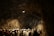 A group of people wearing hard hats are gathered inside a dimly lit cave or tunnel. The rocky walls are illuminated with artificial lighting, casting shadows and creating a rugged atmosphere. The crowd appears to be on a tour or exploration.