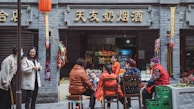 A street scene in front of a small shop with a group of people sitting on chairs and stools conversing. The shop has signs with Chinese characters above its entrance. Several products are visible inside the store, and colorful items are hanging around. A red lantern is hanging from the building, adding a festive touch. The people are dressed casually, with some wearing red clothing.