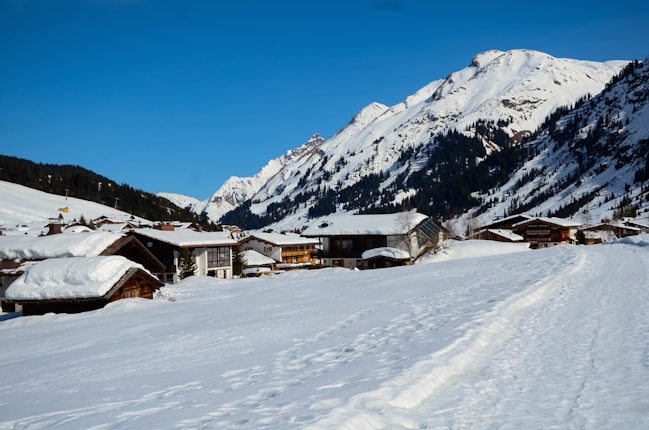 A picturesque alpine village surrounded by snow-covered mountains under a clear blue sky. The wooden houses are blanketed with fresh snow, and the landscape is serene and untouched, with tracks indicating minimal human activity.