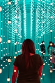 A person stands facing an immersive light installation composed of numerous vertical strands with spherical lights. The lights emit a soft glow in varying shades of teal and orange. The surroundings have a futuristic and serene ambiance, with additional people visible in the background amongst the lights.