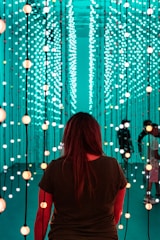 A person stands facing an immersive light installation composed of numerous vertical strands with spherical lights. The lights emit a soft glow in varying shades of teal and orange. The surroundings have a futuristic and serene ambiance, with additional people visible in the background amongst the lights.