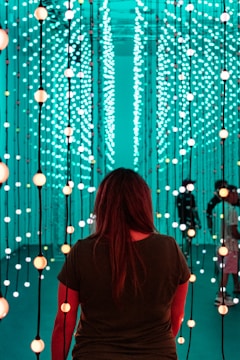A person stands facing an immersive light installation composed of numerous vertical strands with spherical lights. The lights emit a soft glow in varying shades of teal and orange. The surroundings have a futuristic and serene ambiance, with additional people visible in the background amongst the lights.