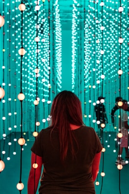 A person stands facing an immersive light installation composed of numerous vertical strands with spherical lights. The lights emit a soft glow in varying shades of teal and orange. The surroundings have a futuristic and serene ambiance, with additional people visible in the background amongst the lights.