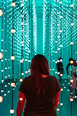 A person stands facing an immersive light installation composed of numerous vertical strands with spherical lights. The lights emit a soft glow in varying shades of teal and orange. The surroundings have a futuristic and serene ambiance, with additional people visible in the background amongst the lights.