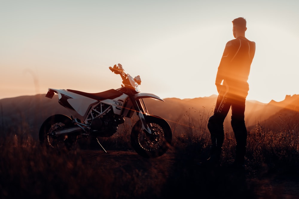 Hombre en chaqueta naranja de pie junto a la motocicleta blanca y negra durante la puesta del sol