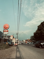 A busy urban street lined with vehicles and shops under a partly cloudy sky. On the left, a sign for 'Cargills Food City' stands prominently. Cars, a small truck, and a tuk-tuk are visible on the road, with power lines overhead stretching into the distance. Trees and buildings surround the area, contributing to the busy yet orderly cityscape.