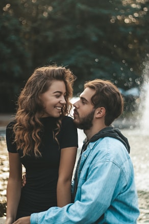 A couple is sitting by a fountain with a blurred forest background. The young woman is smiling warmly while the man gazes at her affectionately. The atmosphere is intimate and serene.