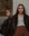 Two young women, both wearing black leather jackets, are positioned against a wall. One is leaning with her back to the wall, hands in her pockets, while the other leans on her side against the wall, looking away. The setting appears to be outdoors, with a blurred natural background.
