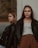 Two young women, both wearing black leather jackets, are positioned against a wall. One is leaning with her back to the wall, hands in her pockets, while the other leans on her side against the wall, looking away. The setting appears to be outdoors, with a blurred natural background.