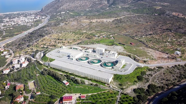 An aerial view of a wastewater treatment facility surrounded by various landscapes, including cultivated fields and a small cluster of residential buildings. The facility features circular water treatment pools and is situated near a road that curves through the area. A hilly terrain extends in the distance with the coastline visible at the horizon.