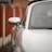 A close-up perspective of a sleek side mirror on a silver car, highlighting its smooth metallic finish and modern design. The background features a blurred red fence, adding contrast to the sharp details of the vehicle.