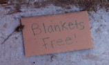 A cardboard sign lying on a concrete surface with the handwritten message 'Blankets Free!'. The surroundings have some dirt and small twigs scattered around the sign.