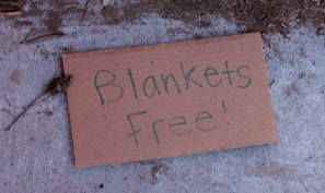 A cardboard sign lying on a concrete surface with the handwritten message 'Blankets Free!'. The surroundings have some dirt and small twigs scattered around the sign.