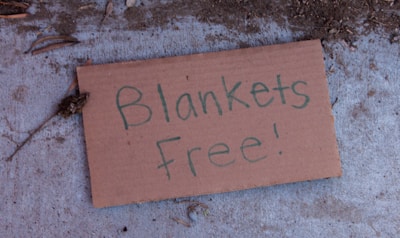 A cardboard sign lying on a concrete surface with the handwritten message 'Blankets Free!'. The surroundings have some dirt and small twigs scattered around the sign.
