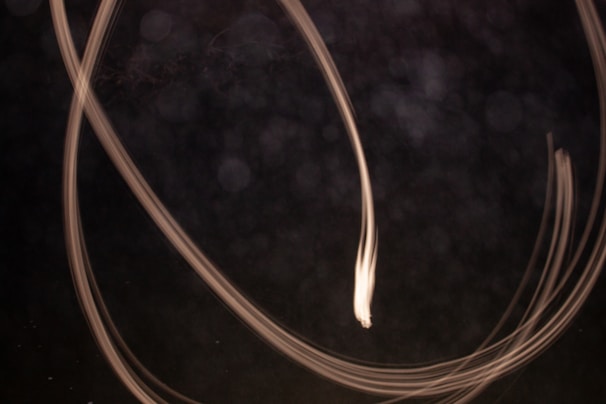 Long exposure photograph capturing light trails against a dark background. The trails curve and twist, creating an abstract and dynamic pattern. The image has a sense of motion and fluidity.