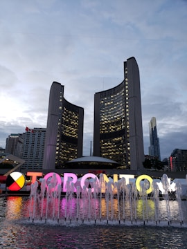 A modern urban scene featuring two curved high-rise buildings against a cloudy sky. In the foreground, a large colorful TORONTO sign is illuminated, accompanied by a series of water fountains. Surrounding the area are additional skyscrapers with lights starting to turn on as evening approaches.