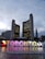 A modern urban scene featuring two curved high-rise buildings against a cloudy sky. In the foreground, a large colorful TORONTO sign is illuminated, accompanied by a series of water fountains. Surrounding the area are additional skyscrapers with lights starting to turn on as evening approaches.