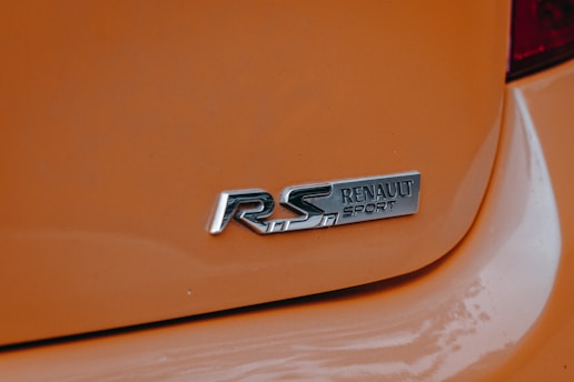 A close-up view of a car's rear badge featuring the 'Renault Sport' logo on an orange background. The metallic lettering stands out against the glossy paint, emphasizing the sporty nature of the vehicle.
