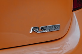 A close-up view of a car's rear badge featuring the 'Renault Sport' logo on an orange background. The metallic lettering stands out against the glossy paint, emphasizing the sporty nature of the vehicle.