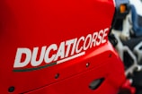A close-up of a bright red motorcycle with the word 'DUCATICORSE' prominently displayed in bold white lettering. The image captures the sleek lines and bold design of the motorcycle's bodywork.