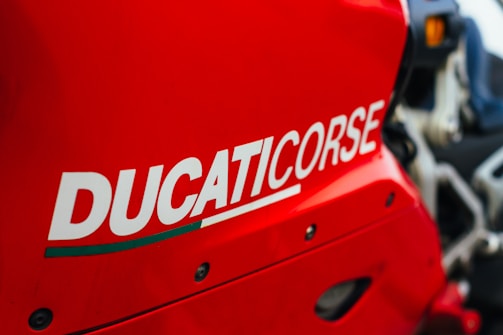 A close-up of a bright red motorcycle with the word 'DUCATICORSE' prominently displayed in bold white lettering. The image captures the sleek lines and bold design of the motorcycle's bodywork.