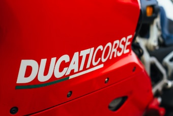 A close-up of a bright red motorcycle with the word 'DUCATICORSE' prominently displayed in bold white lettering. The image captures the sleek lines and bold design of the motorcycle's bodywork.