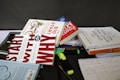 A collection of books and stationery are scattered on a tabletop. The books include titles by authors like Robin Sharma, Simon Sinek, and others. Highlighters and pens are also present alongside a spiral notebook with handwritten notes. The setting appears to be informal, possibly a workspace or study area.