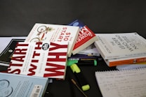 A collection of books and stationery are scattered on a tabletop. The books include titles by authors like Robin Sharma, Simon Sinek, and others. Highlighters and pens are also present alongside a spiral notebook with handwritten notes. The setting appears to be informal, possibly a workspace or study area.