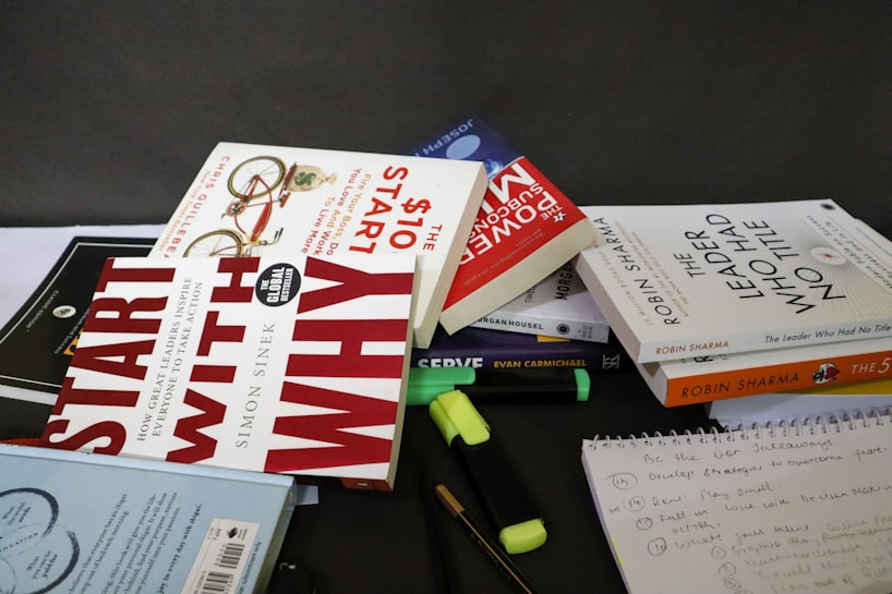 A collection of books and stationery are scattered on a tabletop. The books include titles by authors like Robin Sharma, Simon Sinek, and others. Highlighters and pens are also present alongside a spiral notebook with handwritten notes. The setting appears to be informal, possibly a workspace or study area.