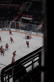 Hockey players dressed in red uniforms are practicing on an indoor ice rink. The rink is surrounded by empty stadium seating and features visible advertisements on the clear barriers. A few players are gathered near the goal post, with one player actively defending it. The atmosphere suggests a quiet, practice session with no audience present.