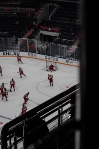 Hockey players dressed in red uniforms are practicing on an indoor ice rink. The rink is surrounded by empty stadium seating and features visible advertisements on the clear barriers. A few players are gathered near the goal post, with one player actively defending it. The atmosphere suggests a quiet, practice session with no audience present.
