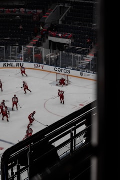 Hockey players dressed in red uniforms are practicing on an indoor ice rink. The rink is surrounded by empty stadium seating and features visible advertisements on the clear barriers. A few players are gathered near the goal post, with one player actively defending it. The atmosphere suggests a quiet, practice session with no audience present.