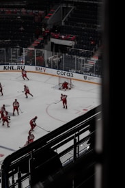 Hockey players dressed in red uniforms are practicing on an indoor ice rink. The rink is surrounded by empty stadium seating and features visible advertisements on the clear barriers. A few players are gathered near the goal post, with one player actively defending it. The atmosphere suggests a quiet, practice session with no audience present.