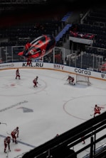 An indoor hockey game in progress with players in red uniforms moving across the ice. The game takes place in a large, modern arena with mostly empty stands. A large blimp featuring advertisements hovers over the arena, and there are visible logos and signage around the rink.