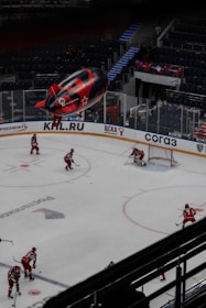 An indoor hockey game in progress with players in red uniforms moving across the ice. The game takes place in a large, modern arena with mostly empty stands. A large blimp featuring advertisements hovers over the arena, and there are visible logos and signage around the rink.