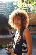 A person with curly hair stands in a well-lit outdoor setting, wearing a sleeveless shirt with graphic prints. The background features wooden structures, greenery, and a couple of people seated, some using electronic devices.