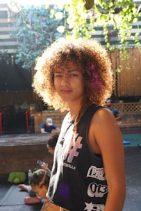 A person with curly hair stands in a well-lit outdoor setting, wearing a sleeveless shirt with graphic prints. The background features wooden structures, greenery, and a couple of people seated, some using electronic devices.