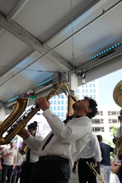 A musician passionately plays a saxophone indoors, surrounded by other musicians in a lively atmosphere. The scene includes a diverse group of people in semi-formal attire enjoying the performance. The background features metal beams and a partial view of a cityscape through large windows.