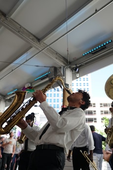 A musician passionately plays a saxophone indoors, surrounded by other musicians in a lively atmosphere. The scene includes a diverse group of people in semi-formal attire enjoying the performance. The background features metal beams and a partial view of a cityscape through large windows.