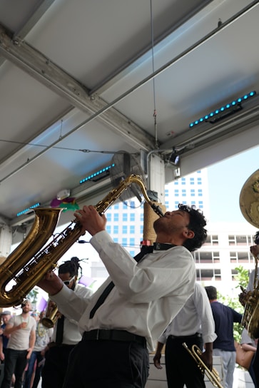 A musician passionately plays a saxophone indoors, surrounded by other musicians in a lively atmosphere. The scene includes a diverse group of people in semi-formal attire enjoying the performance. The background features metal beams and a partial view of a cityscape through large windows.