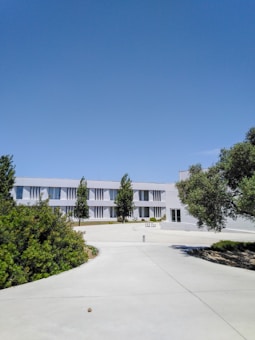 A modern two-story building with a minimalist design, featuring clean lines and large windows. The building is surrounded by well-maintained greenery and a paved walkway leading up to the entrance. Several trees are planted symmetrically in front of the building. The sky is clear and bright blue, contributing to a serene and peaceful atmosphere.