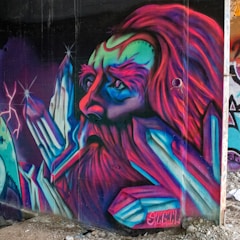 A vibrant graffiti mural depicts a surreal and mystical face surrounded by geometric crystal-like structures. The face is exaggerated with vivid colors of magenta, blue, and green, giving it a fantastical appearance. The details are intricate, with bold lines and a sense of movement created by the flowing hair-like texture.