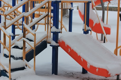 A snowy playground with colorful equipment, including a slide and stairs, covered in white snow. The structures are mostly blue and orange with yellow railings, and the ground is blanketed in snow.