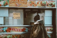 A person stands in front of a food truck with a sign that says 'Only Cash.' The truck advertises various kinds of halal food items such as chicken over rice, lamb over rice, and falafel over rice. The person is wearing a warm jacket and a beanie, suggesting a cold environment, and the truck's images and text are vibrant and colorful.
