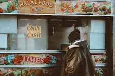 A person stands in front of a food truck with a sign that says 'Only Cash.' The truck advertises various kinds of halal food items such as chicken over rice, lamb over rice, and falafel over rice. The person is wearing a warm jacket and a beanie, suggesting a cold environment, and the truck's images and text are vibrant and colorful.