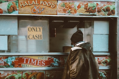 A person stands in front of a food truck with a sign that says 'Only Cash.' The truck advertises various kinds of halal food items such as chicken over rice, lamb over rice, and falafel over rice. The person is wearing a warm jacket and a beanie, suggesting a cold environment, and the truck's images and text are vibrant and colorful.